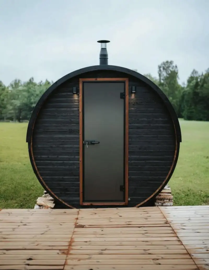 Sauna tonneau en bois noir avec porte vitrée et cheminée, dans un pré