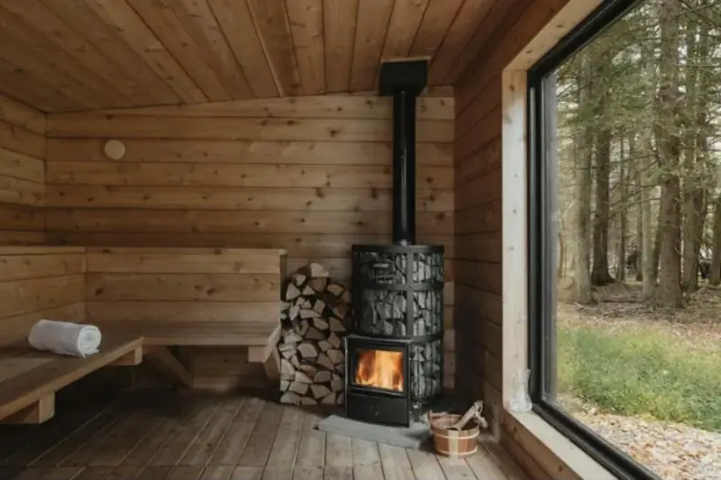 Intérieur de sauna en bois avec poêle à bois allumé et grande baie sur la forêt